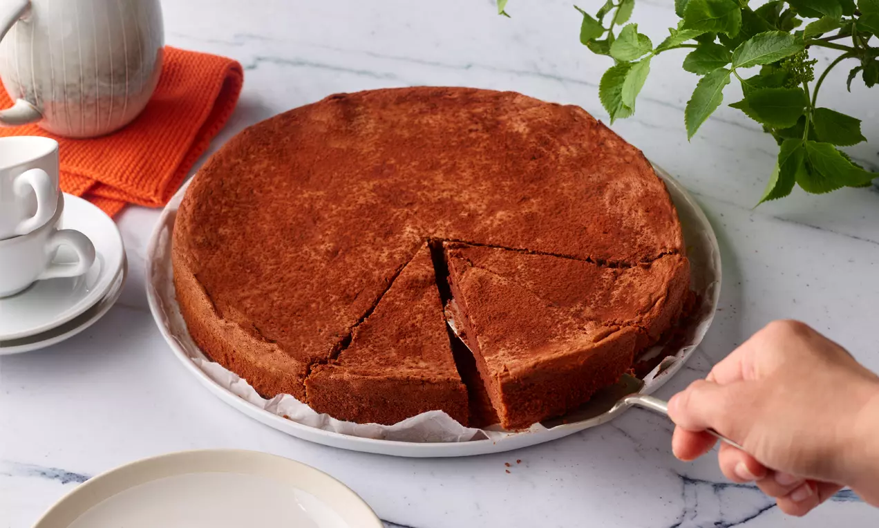 Schokoladen-Maronikuchen mit glatter, dunkelbrauner Oberfläche, angeschnitten, auf weißem Teller mit Papier, daneben Tasse und grüne Pflanze