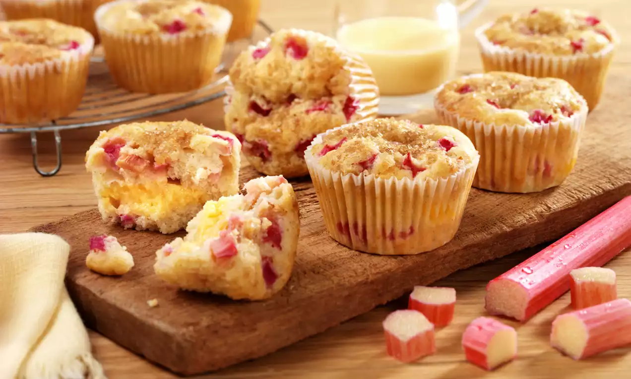 Golden muffins dotted with soft red rhubarb chunks and creamy custard, topped with a crumbly golden brown finish on a wooden board