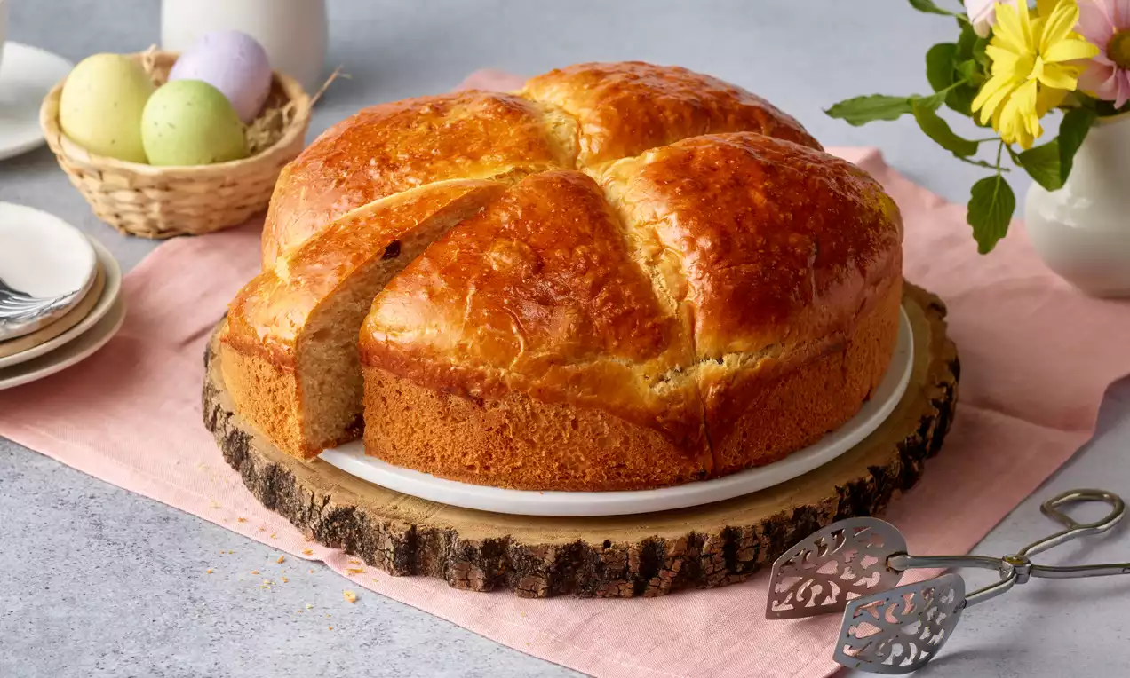 Osterpinze mit goldbrauner Kruste, rund und leicht eingeschnitten, auf einem rustikalen Holzbrett mit rosa Tischdecke und Osterdeko