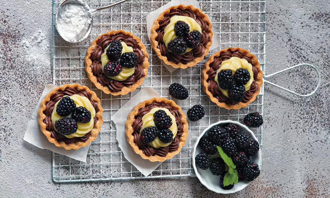 Krehké tartaletky s mierne zlatistým okrajom, plnené žltým krémom a čokoládovou ozdobou, dozdobené čerstvými černicami na mriežke.