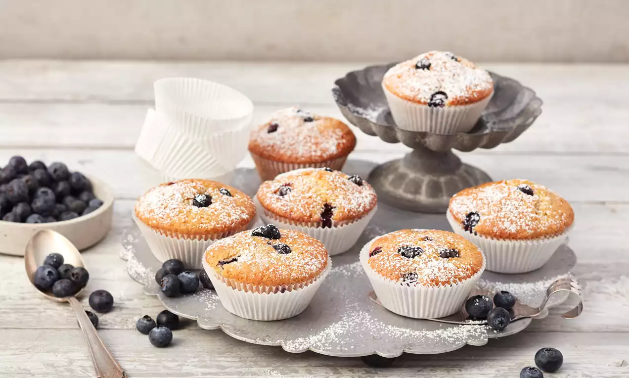 Goldbraune Blaubeer-Muffins mit Puderzucker bestäubt in weißen Papierförmchen auf silbernem Teller, daneben frische Blaubeeren und Löffel