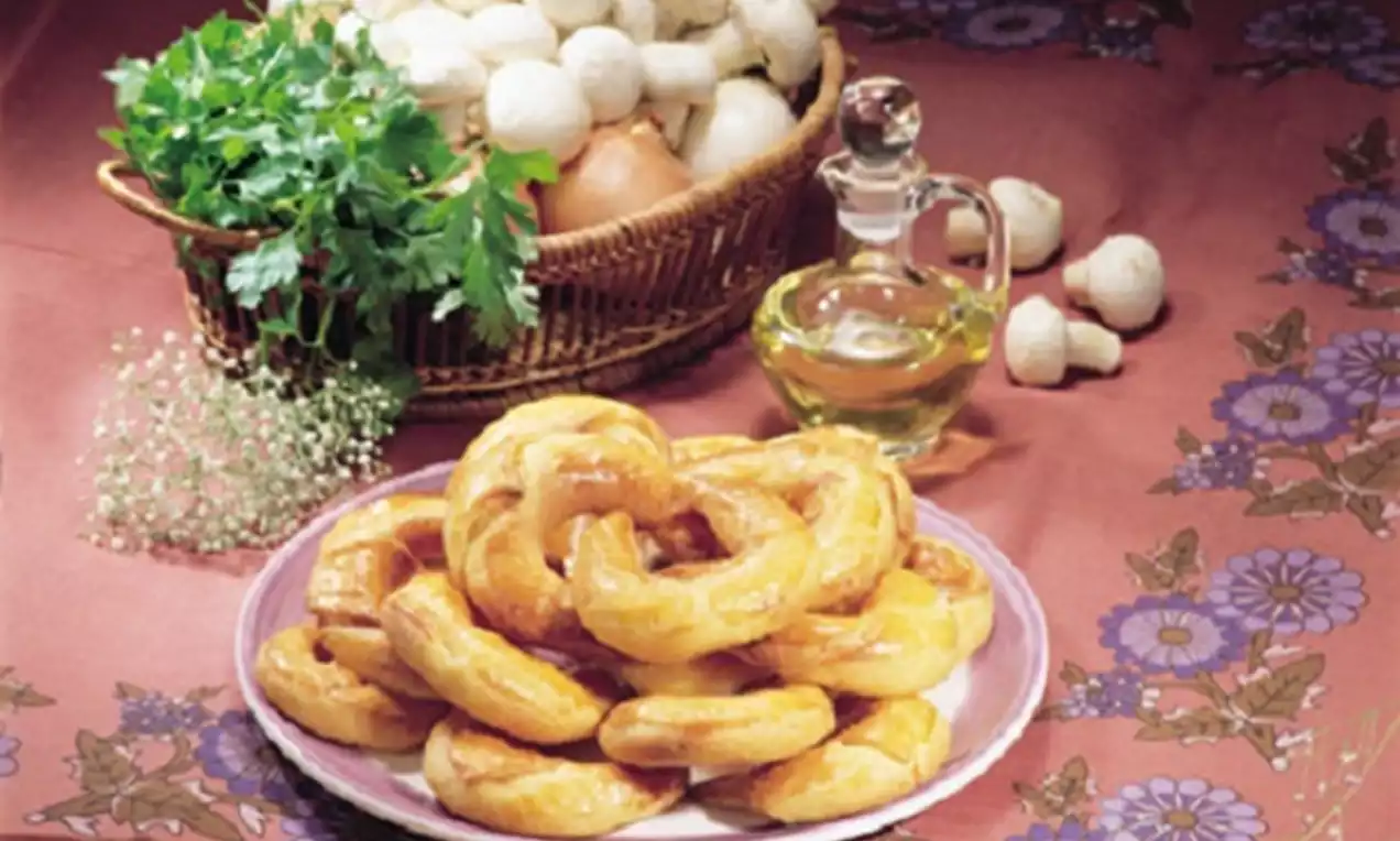 Altın rengi ay çörekleri beyaz bir tabakta sıralı, arka planda bir sepet dolusu mantar, taze yeşillik ve zeytinyağı şişesi yer alıyor