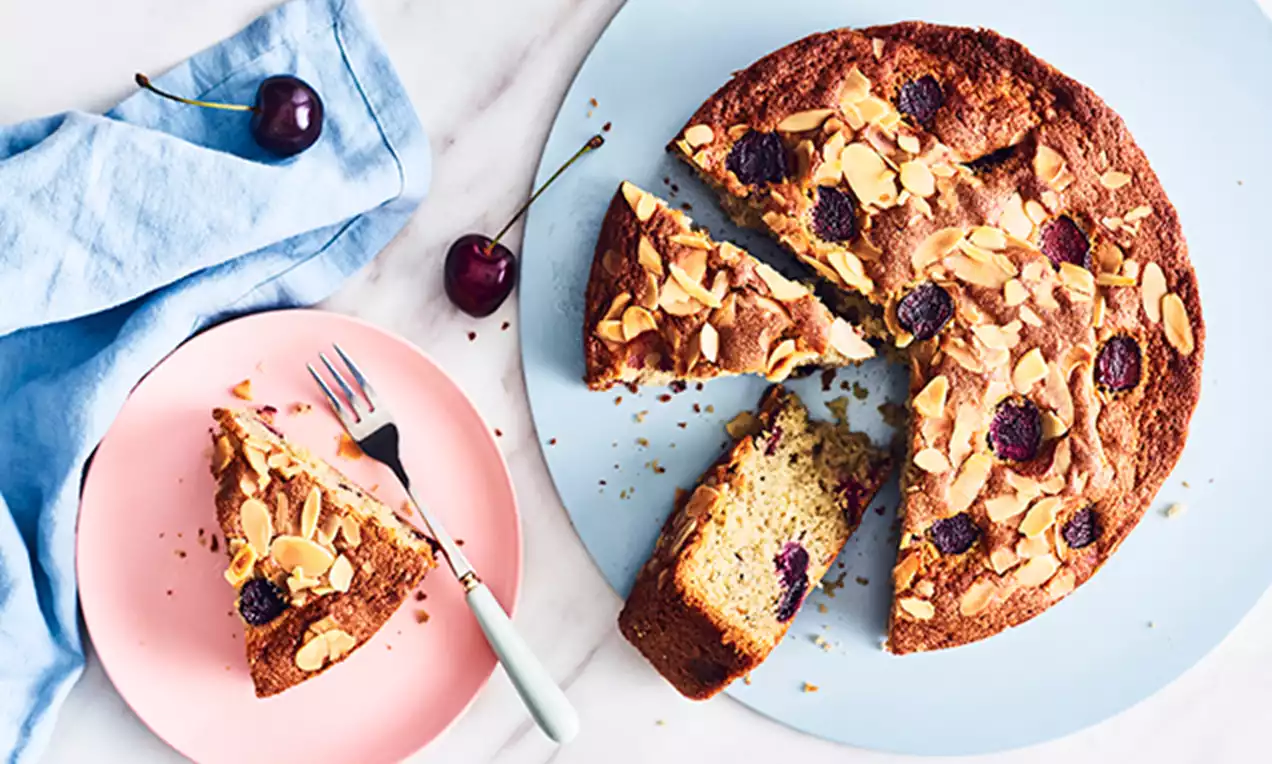 Golden-brown cherry cake with almond flakes, featuring whole cherries baked in, sliced on pastel plates with fresh cherries alongside