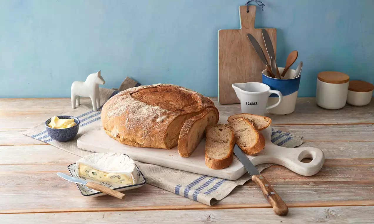 Walnussbrot mit goldbrauner, rissiger Kruste auf einem weißen Holzbrett, umgeben von geschnittenen Scheiben, Käse und Butter