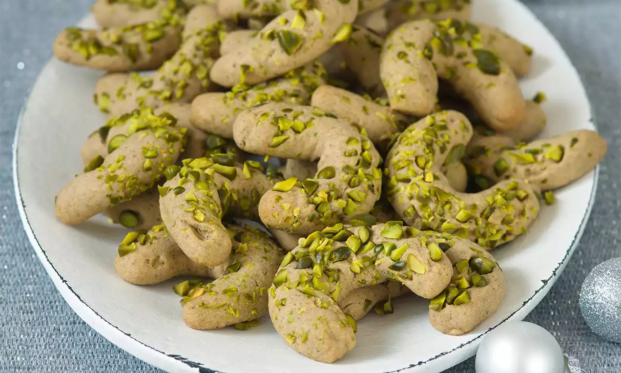 Pistáciové rohlíčky ve tvaru zahnutých rohů, světle hnědé barvy s posypem zelených nasekaných pistácií, na bílém talíři s lesklými ozdobami