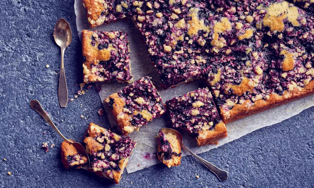 Mustikkapiirakka on leikattu neliskanttisiksi paloiksi, kullankeltaisen pohjan päällä mustikoita ja muruseosta, aseteltu sattumanvaraisesti lusikoiden ympäröimänä