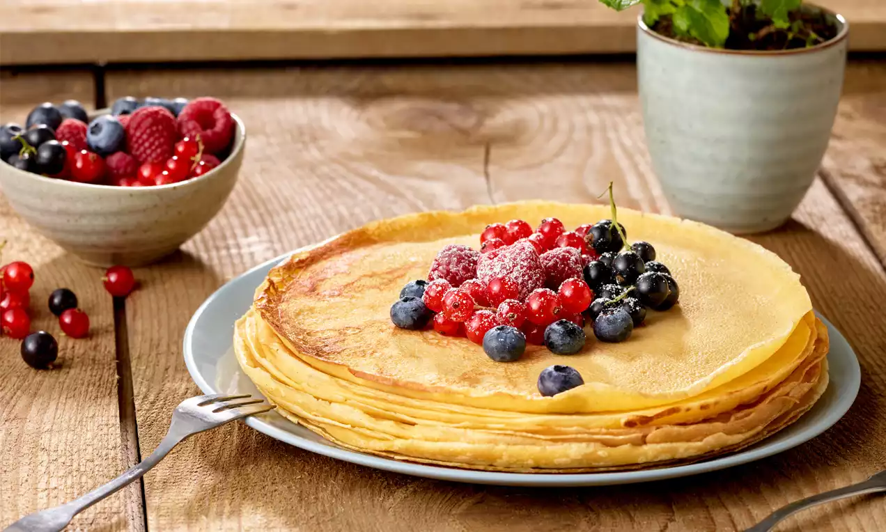 Goldgelbe Palatschinken als gestapelter Stapel auf hellblauem Teller, belegt mit roten und blauen Beeren sowie Puderzucker, daneben eine Schale Beeren
