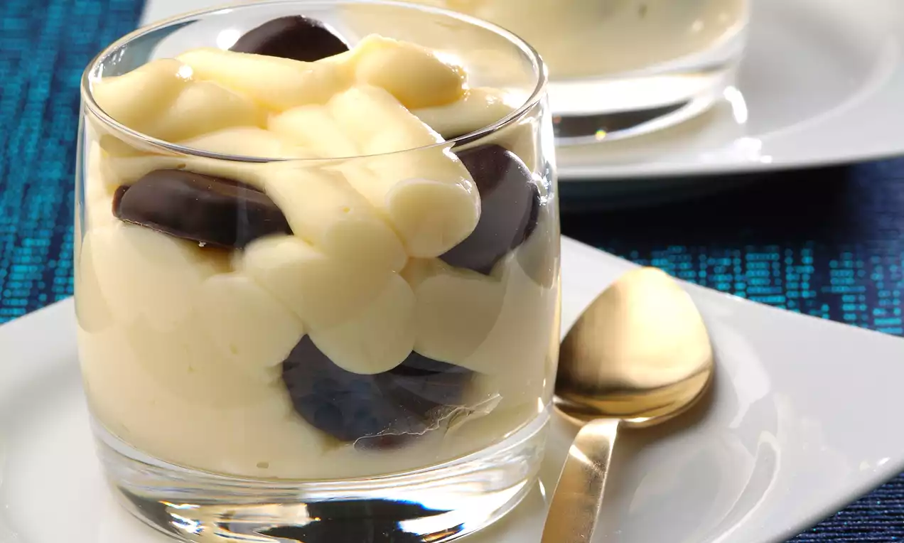 Cam bir bardakta sunulan tatlı, yumuşak sarı muzlu krema katmanları ve koyu çikolata parçalarıyla ışık saçan pürüzsüz bir görünüme sahip