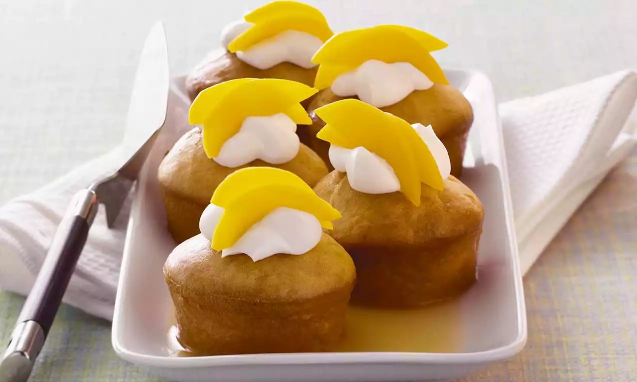 Muffin dorati e soffici imbevuti di sciroppo, decorati con soffici ciuffi di panna bianca e fette di mango giallo intenso su un piatto rettangolare
