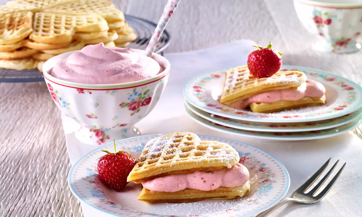 Srcoliki vafli posuti šećerom u prahu, punjeni ružičastom kremom od jagoda, s jagodom na vrhu, na bijelim tanjurima s cvjetnim uzorkom