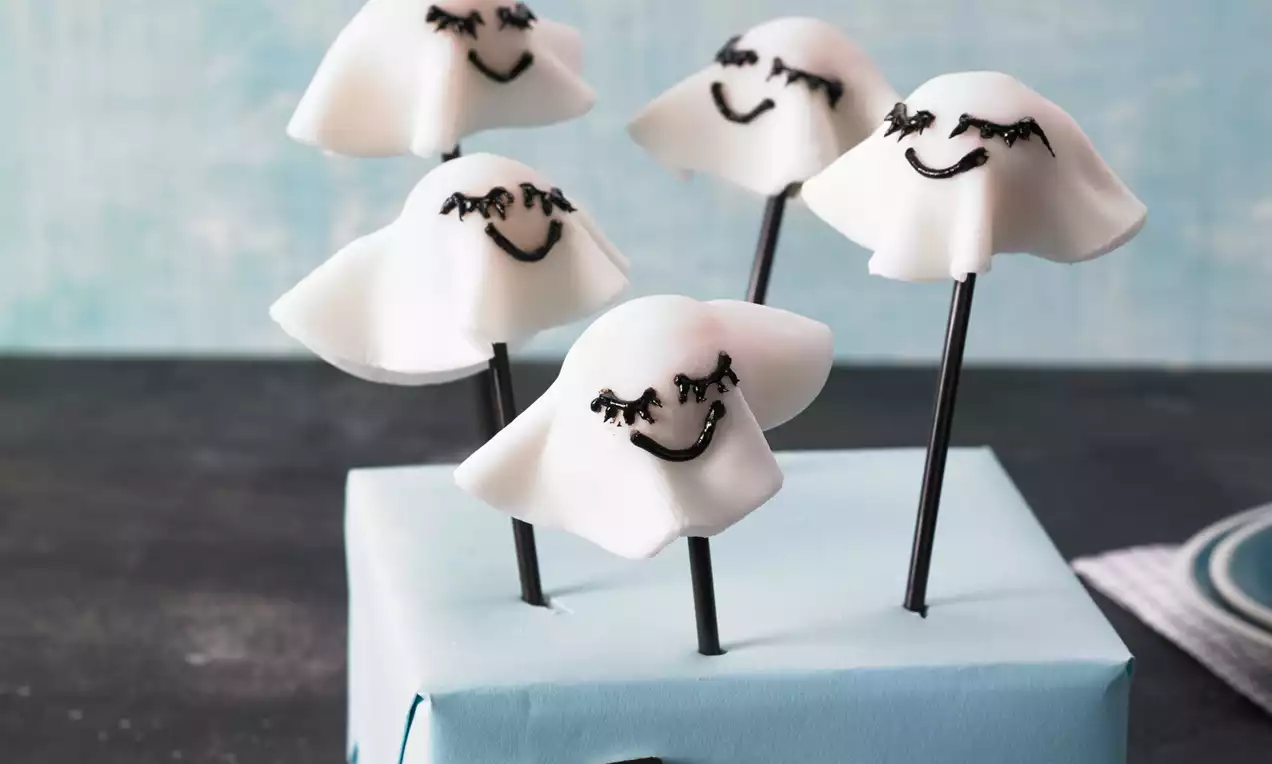 Weiße Halloween-Cake-Pops in Form von Geisterfiguren mit schwarzen Gesichtszügen auf schwarzen Stielen in hellblauem Präsentationsblock
