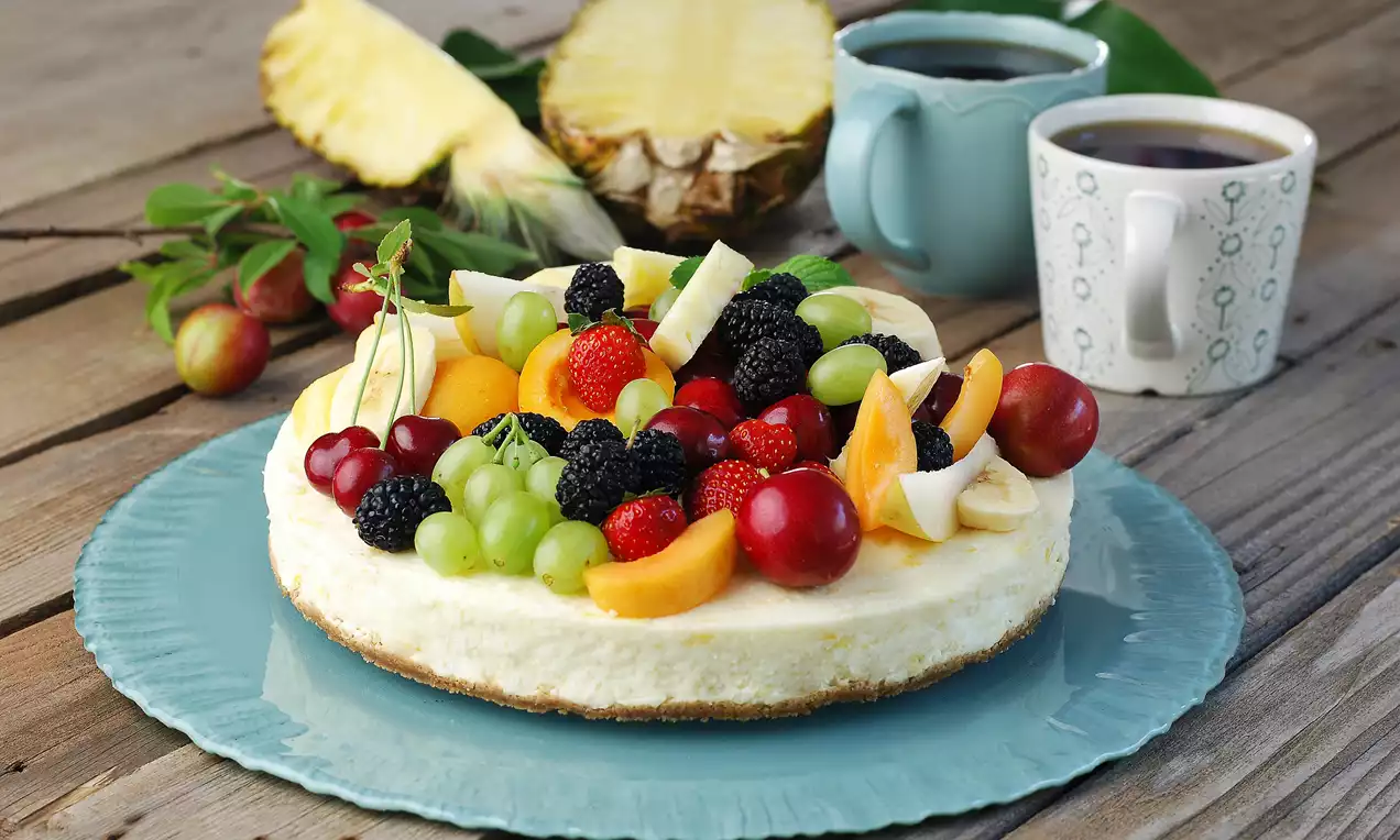 Kremalı tabanı üzerinde çilek, böğürtlen, üzüm, şeftali ve muz dilimleriyle süslenmiş, mavi bir tabakta servis edilen meyveli cheesecake