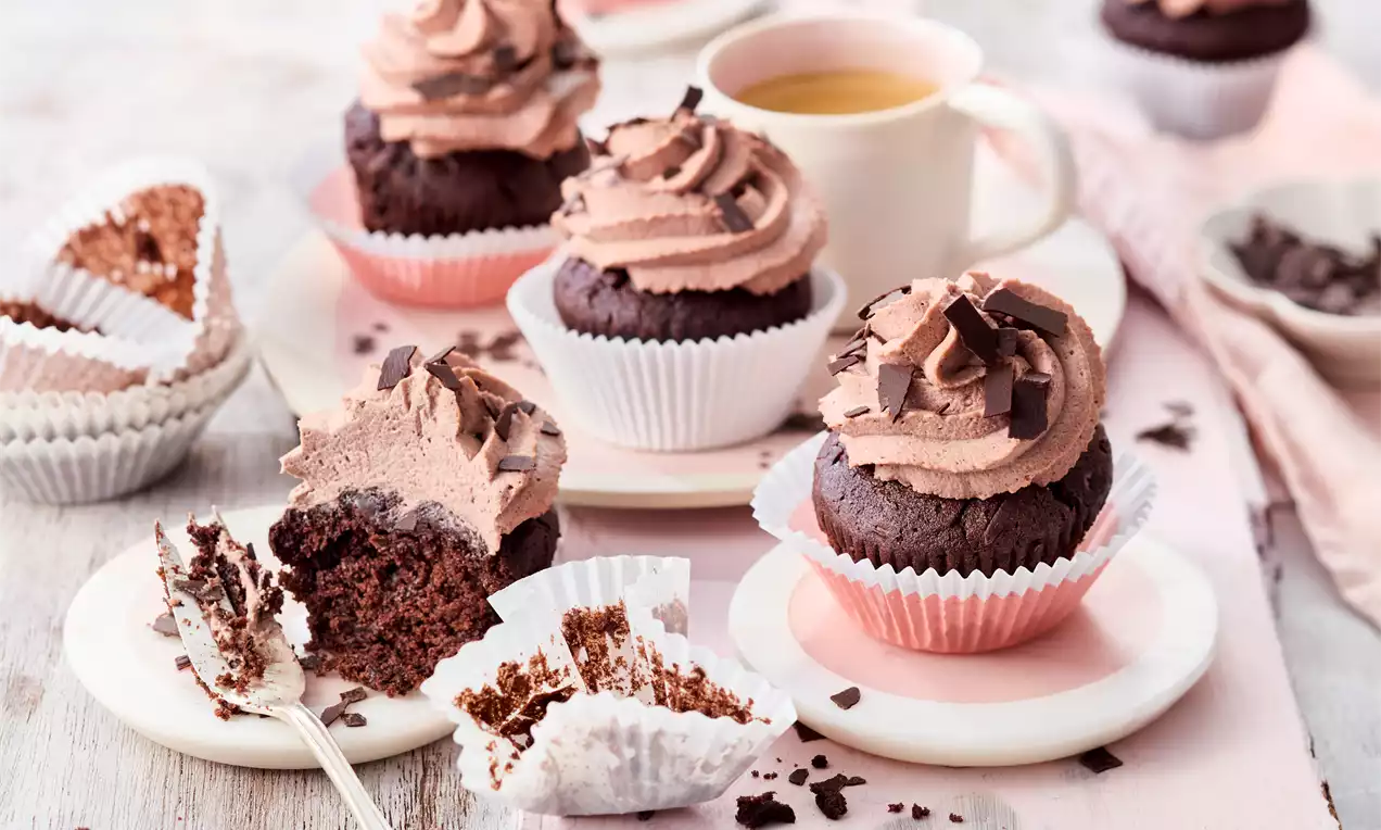 Schoko-Cupcakes mit flauschiger Kakaocreme und Schokoraspeln in Papierförmchen auf Tellern, daneben zerbröselte Förmchen und eine Tasse