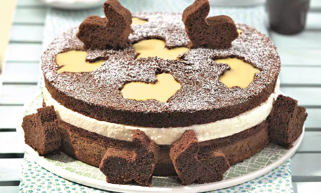 Tort cu blat de cacao presărat cu zahăr pudră, decupaje în formă de iepurași cu cremă galbenă, strat alb de cremă și decorațiuni de iepurași de ciocolată