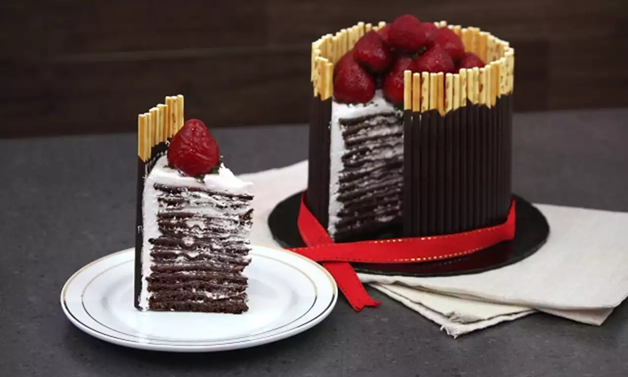Layered chocolate crepe cake with dark cocoa crepes and white cream filling, topped with red raspberries and encircled by chocolate sticks