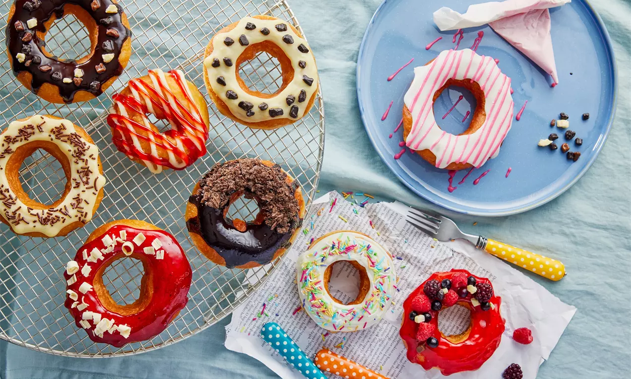 Renkli sırlı ve farklı malzemelerle süslenmiş çeşitli donutlar, tel ızgara ve mavi tabak üzerinde, yanlarında desenli çatal bıçaklarla sunulmuş