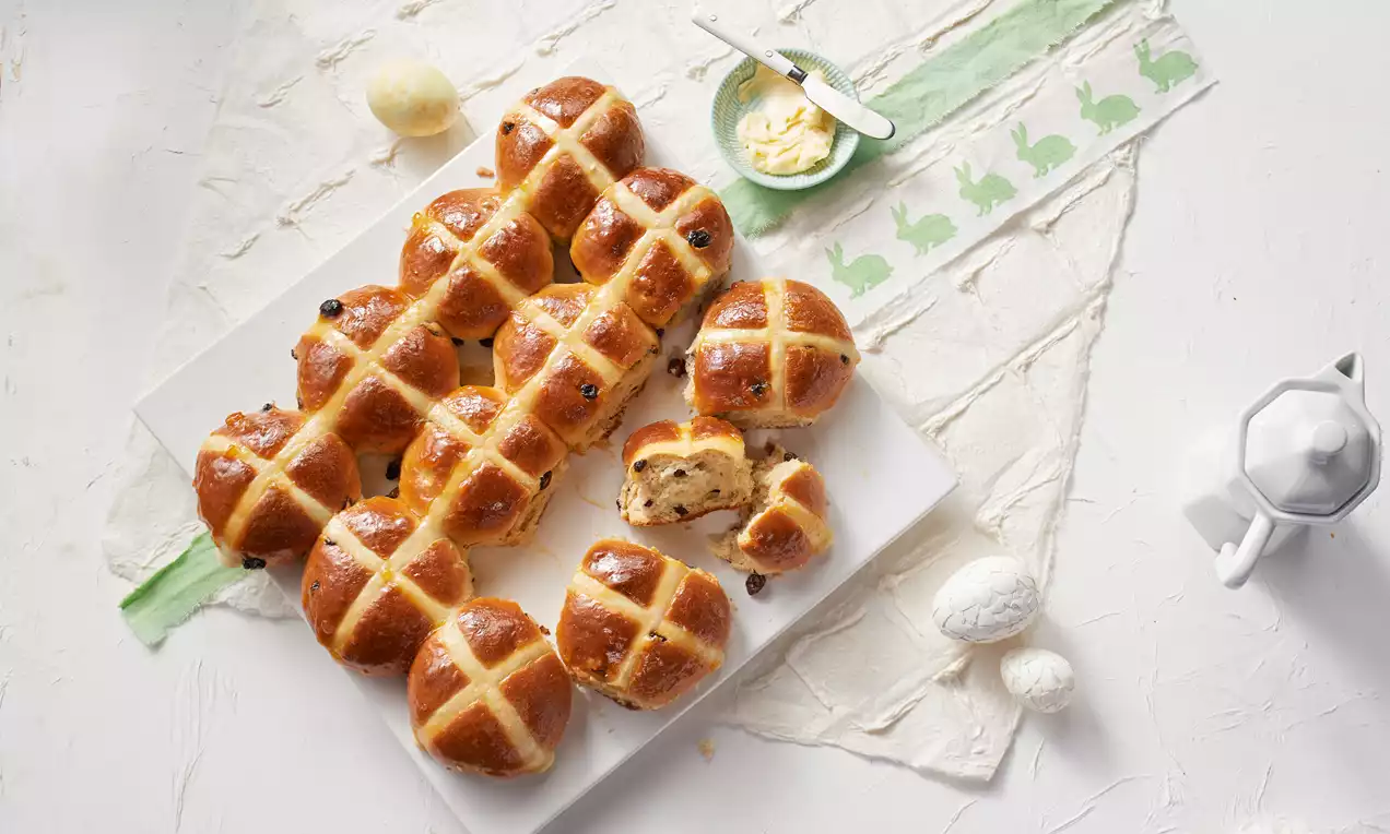 Goldbraune Hot Cross Buns mit glänzender Oberfläche und weißen Kreuzmustern, arrangiert auf weißer Platte neben Butter und Osterdeko