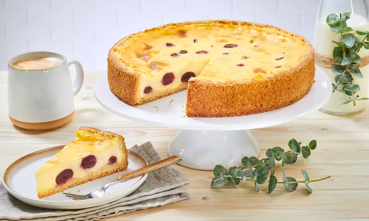 Runder Kirsch-Schmand-Kuchen mit goldbraunem Mürbeteigrand und heller Füllung, aufgeschnitten auf weißer Tortenplatte mit Eukalyptusdeko