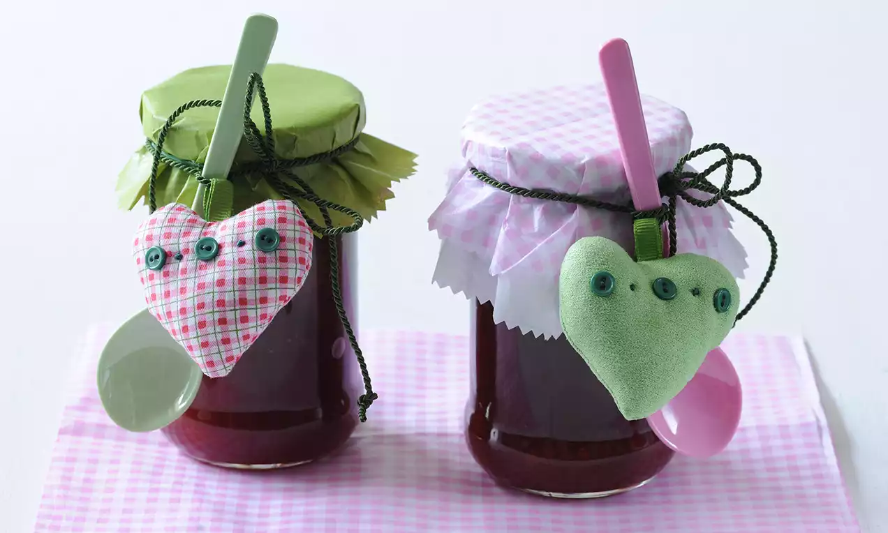 Dva skleněné zavařovací sklenice s jahodovo-rybízovým džemem, dekorované víčky s látkovými potahy v zelené a růžové barvě, připevněné lžičkami a látkovými srdíčky