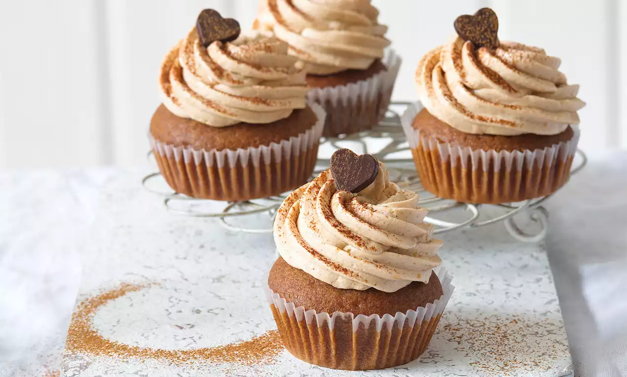 Perníkové cupcakes s hnědým základem, krémovou šlehačkou posypanou skořicí, zdobené čokoládovým srdcem na mřížce a jemném podkladu