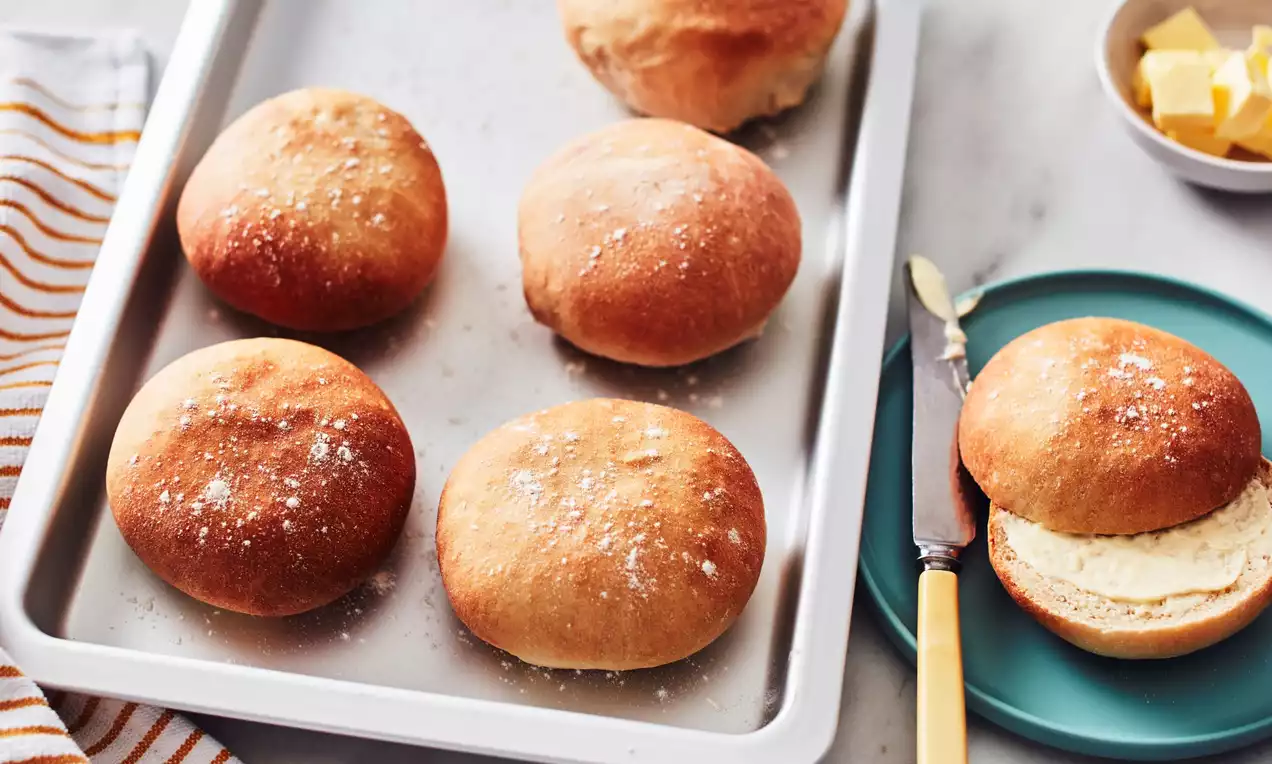 Golden brown round bread rolls with a soft texture, lightly dusted with flour on a metal tray beside a bun on a blue plate with butter