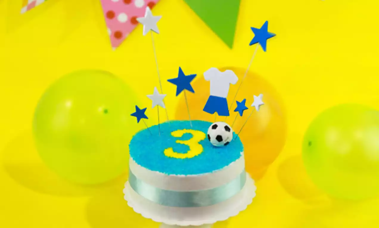 Round sponge cake with smooth white sides wrapped in a light blue ribbon, bright blue top with a yellow number 3, a small soccer ball, blue and white star toppers, and a shirt and shorts decoration on a white plate against a vibrant yellow backdrop with balloons and bunting.