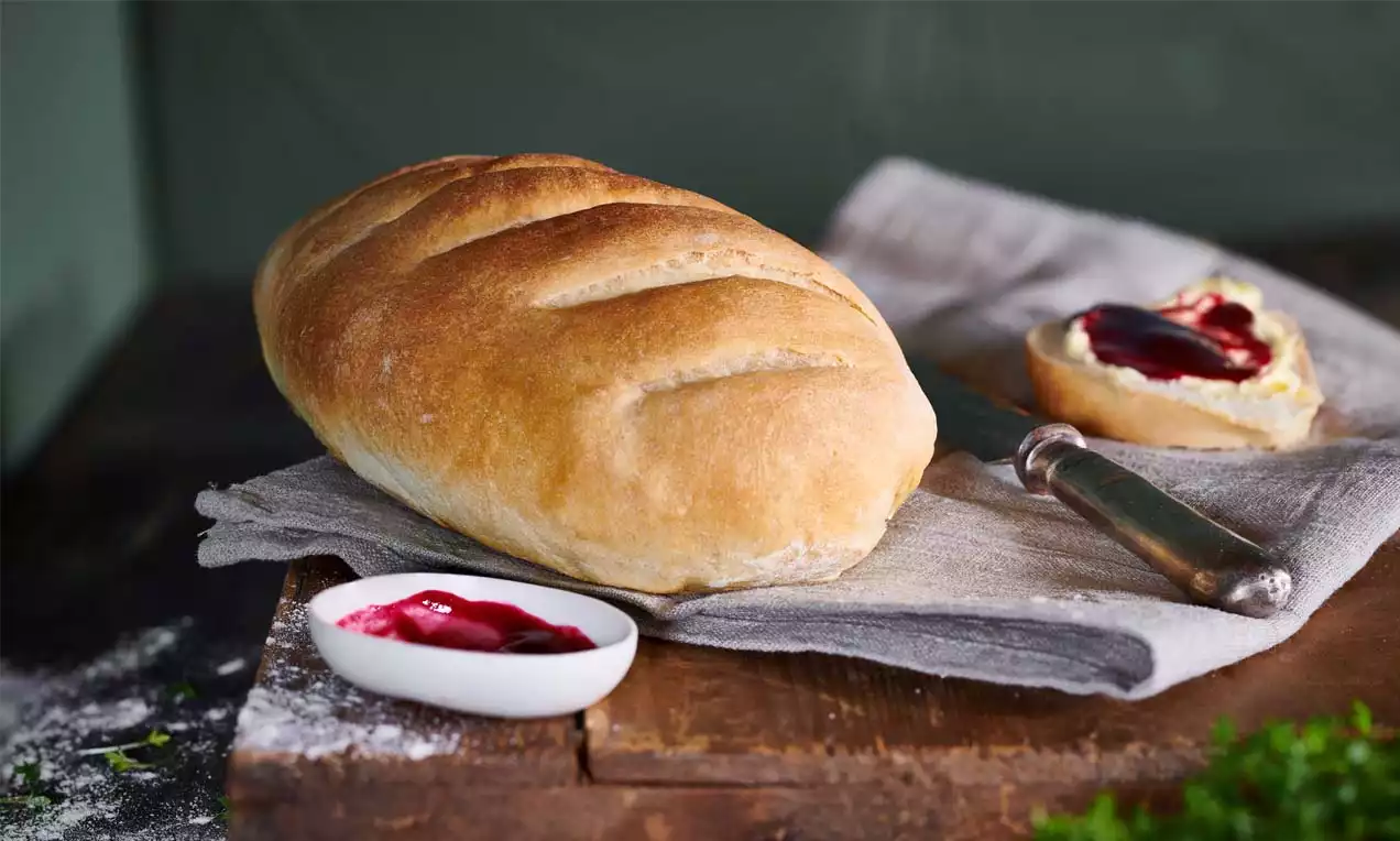 Hellgoldenes, längliches Brot mit glatter, leicht bemehlter Kruste auf grauem Leinentuch, daneben Marmelade und ein bestrichenes Brötchen