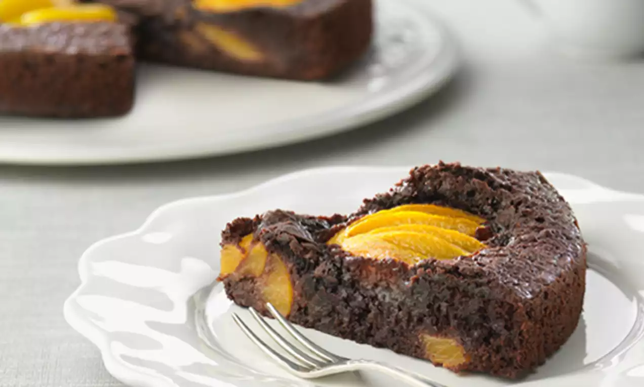 Porción de tarta de chocolate oscuro con rodajas de melocotón amarillo brillantes en el centro sobre un plato blanco con tenedor