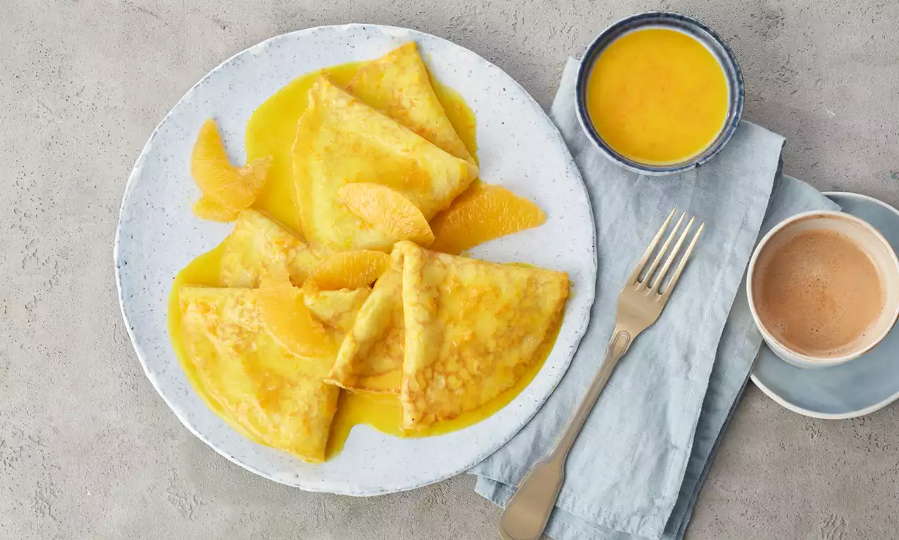 Crepes-Suzette mit goldgelber Sauce und Orangenspalten kunstvoll arrangiert auf einem hellen Teller neben einer Tasse Espresso und Besteck