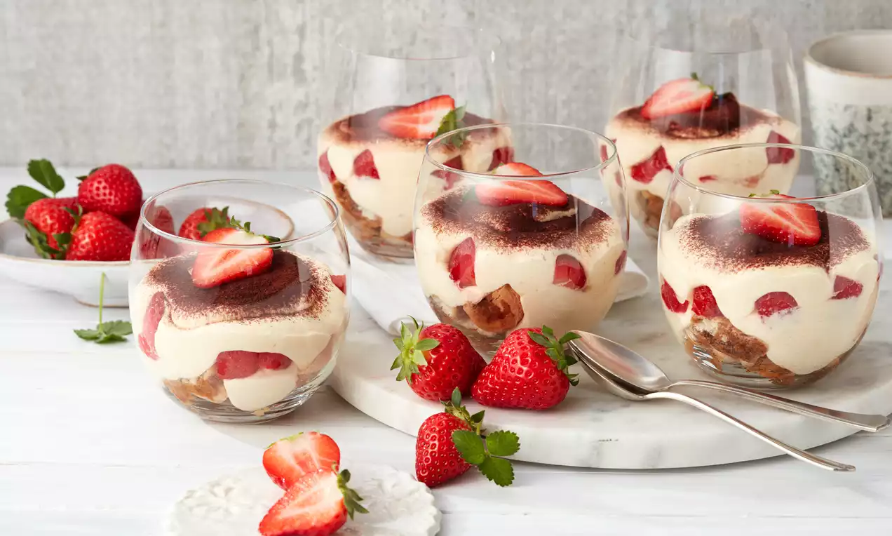 Erdbeer-Tiramisu in klaren Gläsern mit luftiger Creme, roten Erdbeerscheiben, getränktem Biskuit und Kakaopulver auf einer weißen Platte