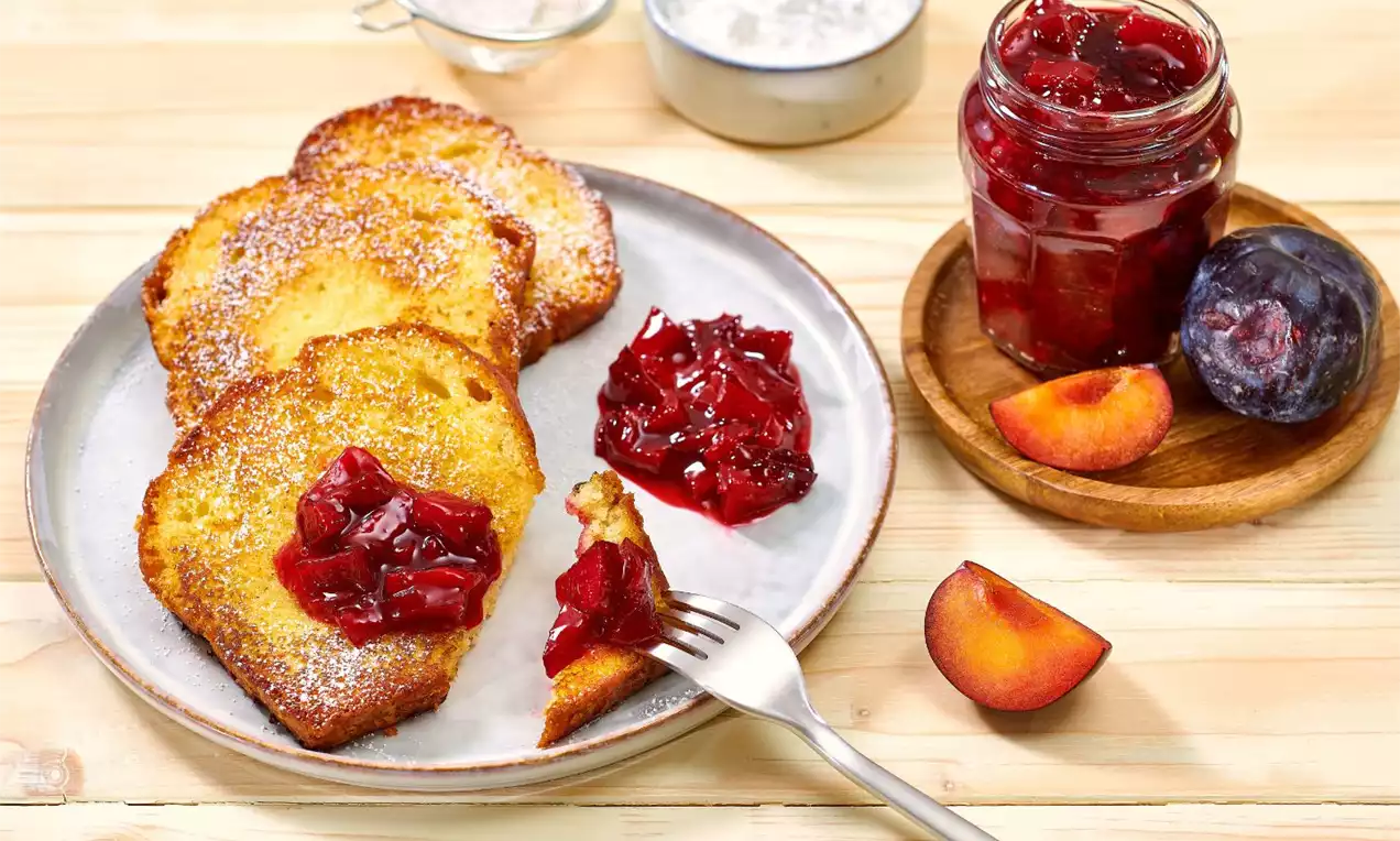 Zlatavé plátky French toastu s křupavým povrchem, zdobené tmavě červenou švestkovou marmeládou na matném šedém talíři s čerstvými švestkami