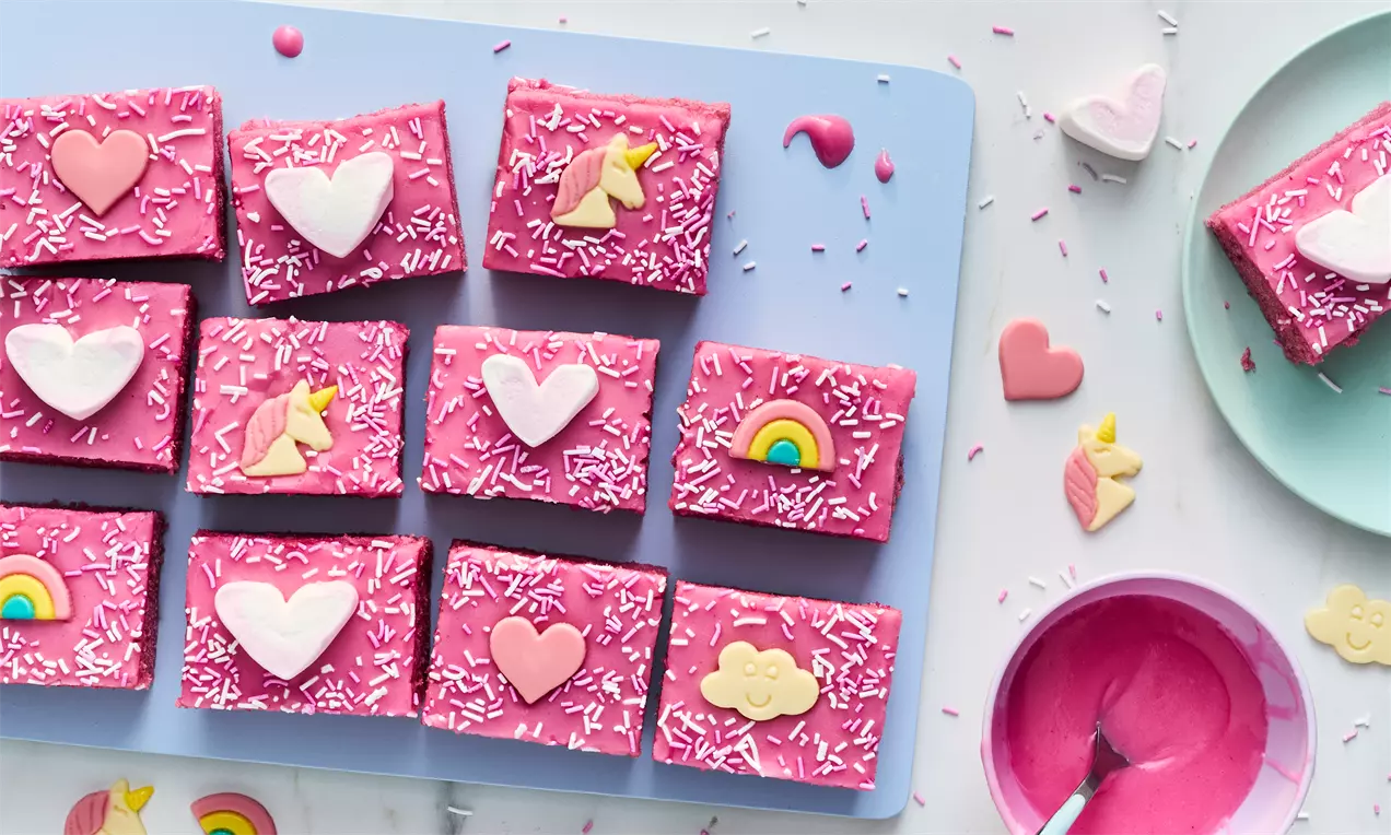 Bright pink unicorn-themed squares topped with pastel decorations like hearts, rainbows, and sprinkles on a pale blue tray