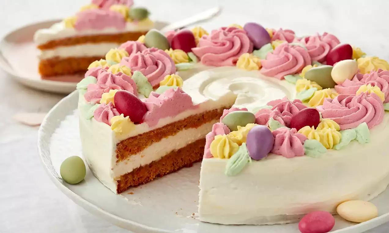Easter cake with white cream frosting, two brown sponge layers, pastel rosettes, candy eggs, and a slice on a plate in the background