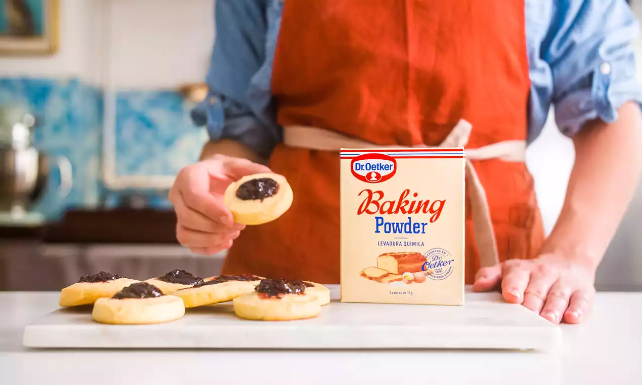 Galletas redondas de base dorada y textura tierna con un centro de mermelada oscura dispuestas sobre una tabla blanca en una cocina