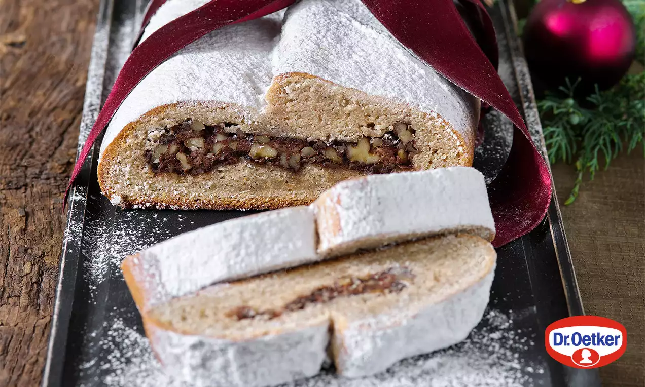 Vánoční štola zlatohnědá, jemně obalená v moučkovém cukru, plněná směsí oříšků a mandlí, na tmavém tácku vedle červené stuhy a vánoční ozdoby