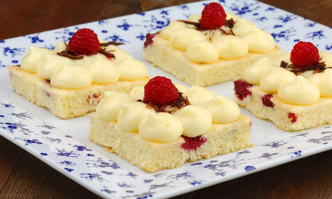 Altın sarısı kek tabanlı kare pastacıklar, üzeri kremalı yumuşak dokulu, ahududu ve çikolata parçalarıyla süslenmiş porselen tabakta