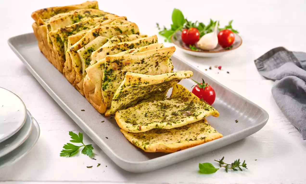Goldbraunes Faltenbrot mit grüner Kräuterbutterfüllung, auf einem rechteckigen Teller angerichtet, dekoriert mit frischen Kräutern und Tomaten
