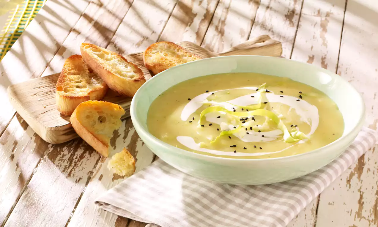 Cremiges Spitzkohlsüppchen in zartgrüner Schale, dekoriert mit Öl und schwarzem Sesam, dazu geröstete Brotscheiben auf Holzbrett und Stofftuch