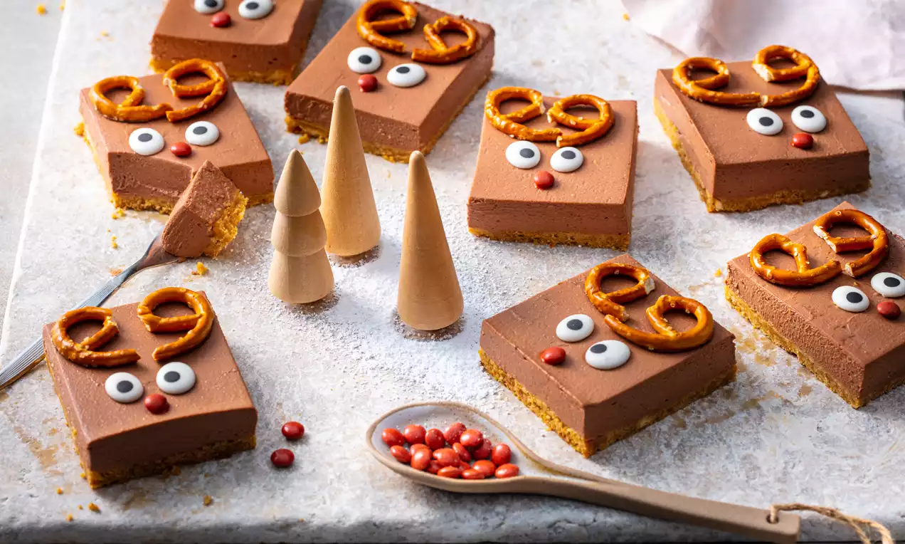 Vierkante no bake cheesecakejes met een gladde bruine topping, versierd met pretzels, witte oogjes en rode neuzen op een licht poedersuikerbed