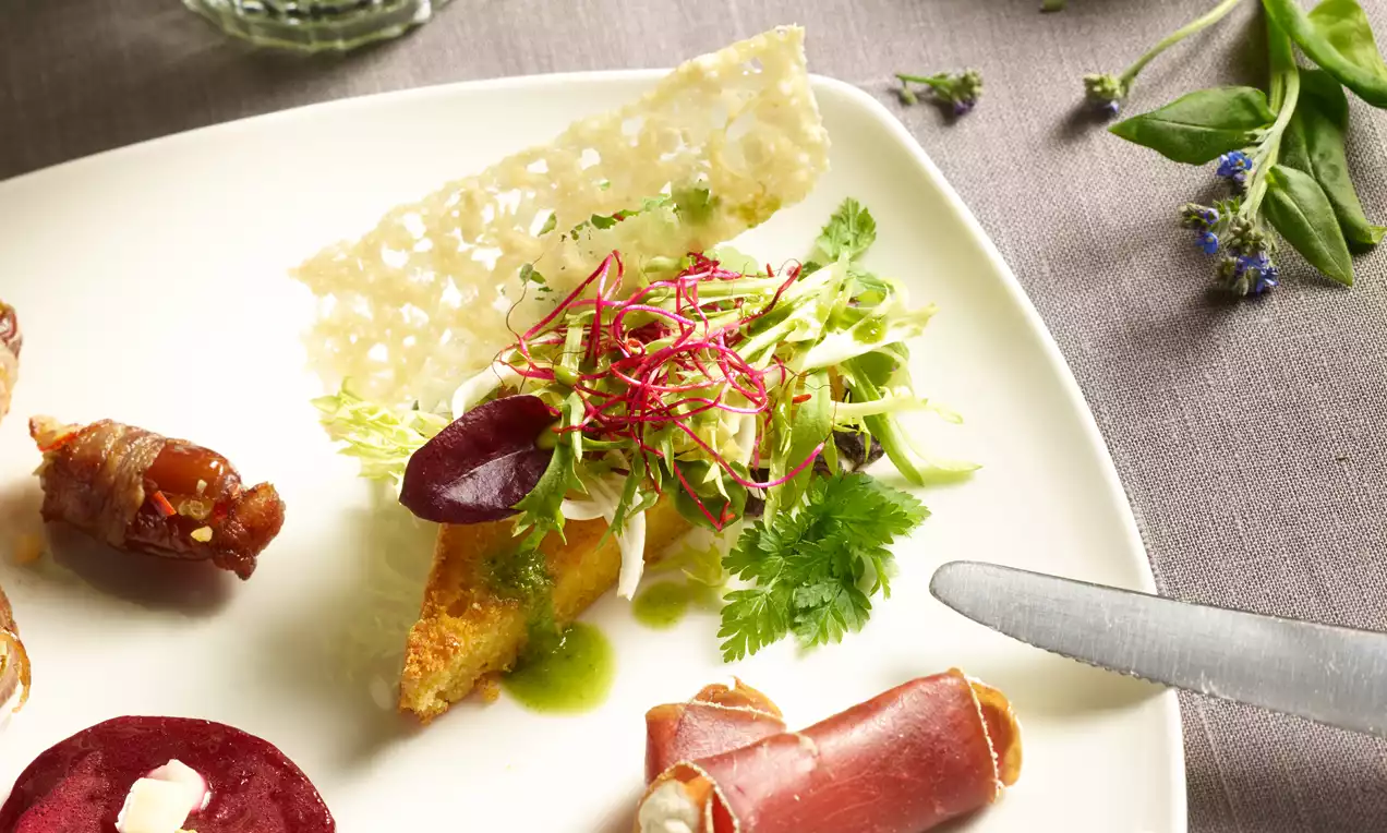 Maischnitten mit goldener Kruste, belegt mit frischem Kräutersalat, roten Sprossen und grüner Sauce auf einem weißen Teller mit Käsestange