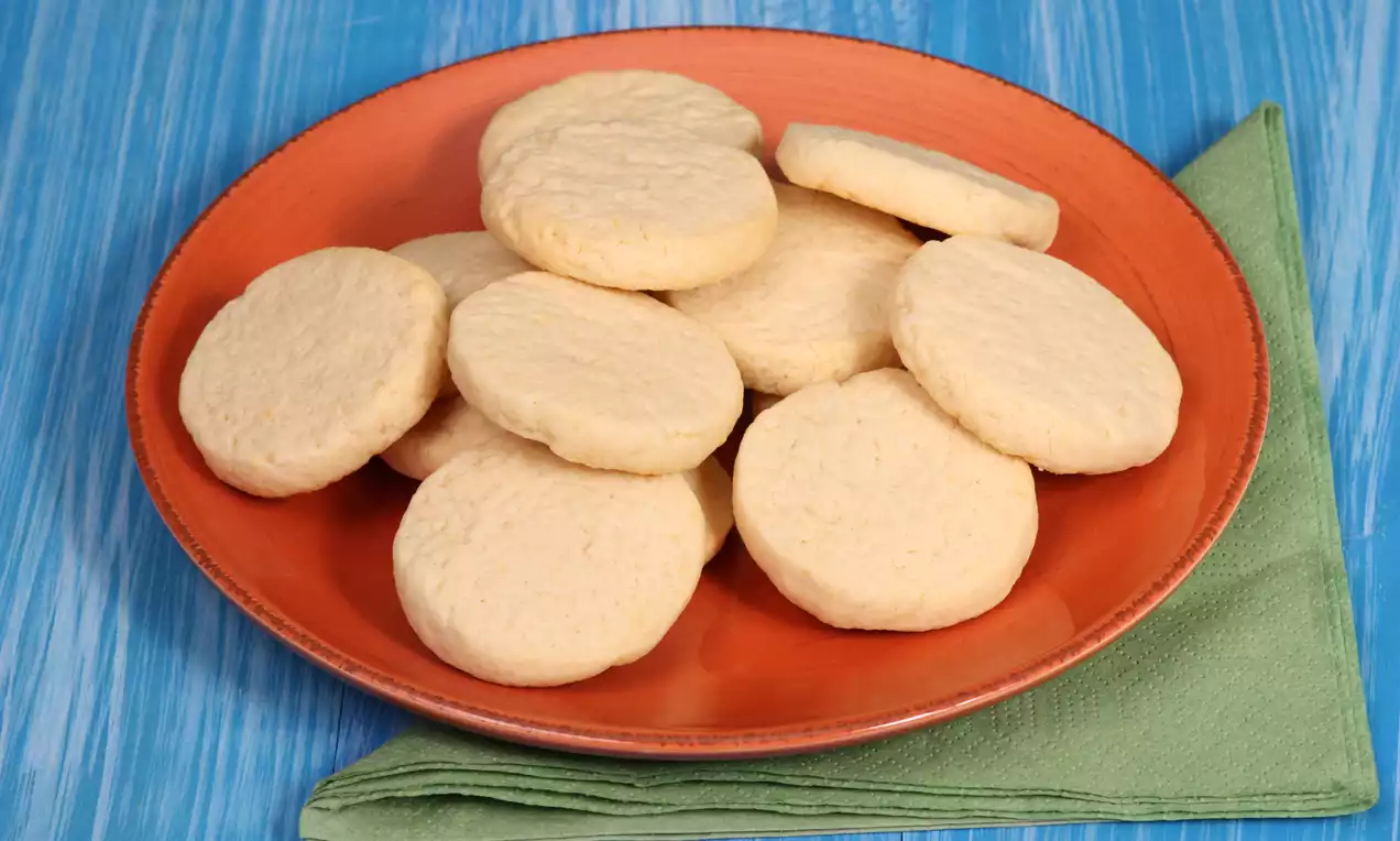 Yuvarlak, açık krem tonlarında kurabiyeler kırmızı tabakta düzenli şekilde yerleştirilmiş ve tabak yeşil bir bez üzerine konmuş