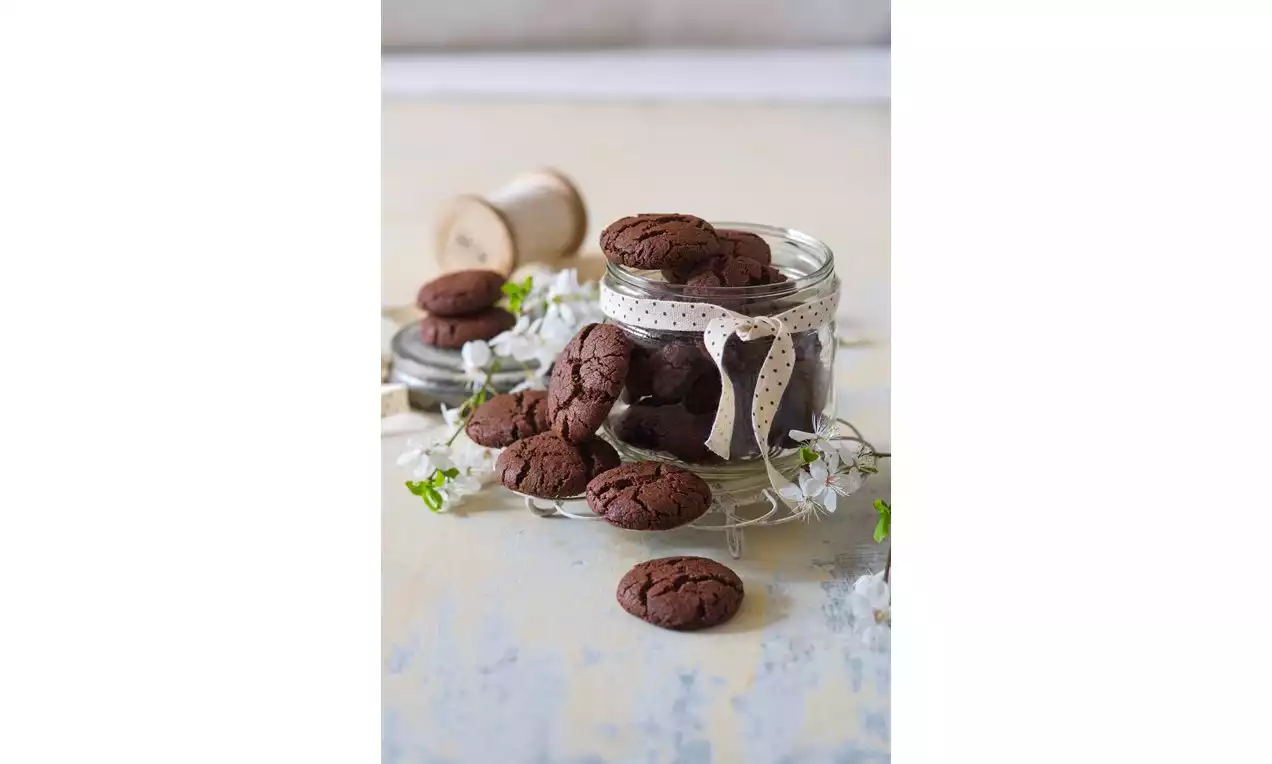 Tmavě hnědé kakaové cookies s popraskanou texturou naskládané v průhledné sklenici s mašlí na světlém povrchu, obklopené bílými kvítky