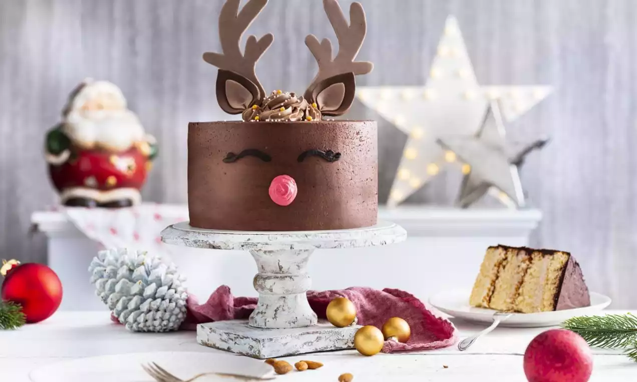 Round chocolate cake with smooth texture, antler-shaped toppers, red fondant nose and gold accents on a white stand surrounded by festive decor