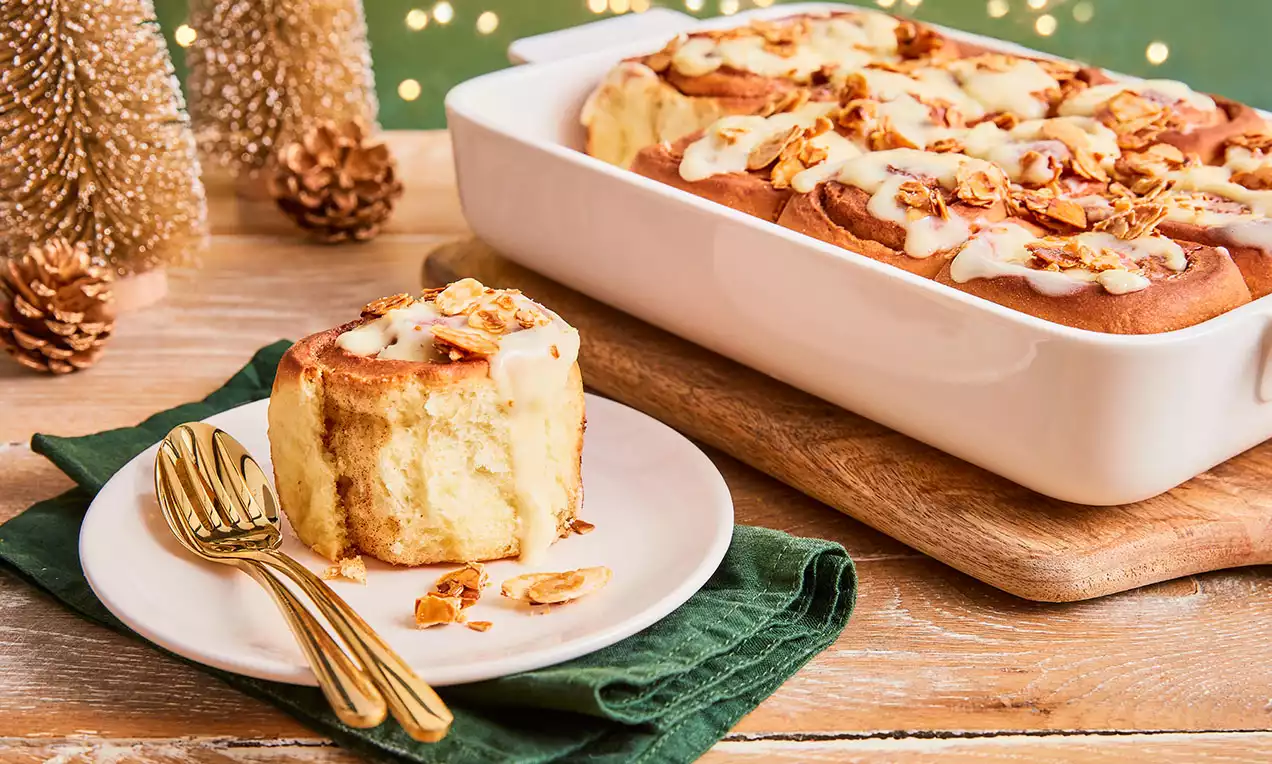 Weihnachtliche Zimtschnecke mit goldbraunem Teig und weißem Frischkäsetopping auf weißem Teller mit goldfarbenem Besteck auf grünem Stoff