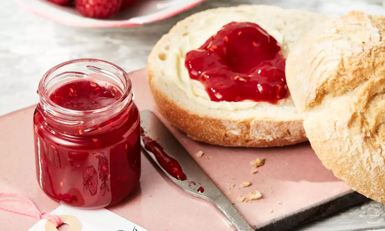 Sytě červený jahodovomalinový džem s jemnou texturou v malém skleněném závitovém skleničce vedle rozříznutého chleba s máslem na růžové podložce