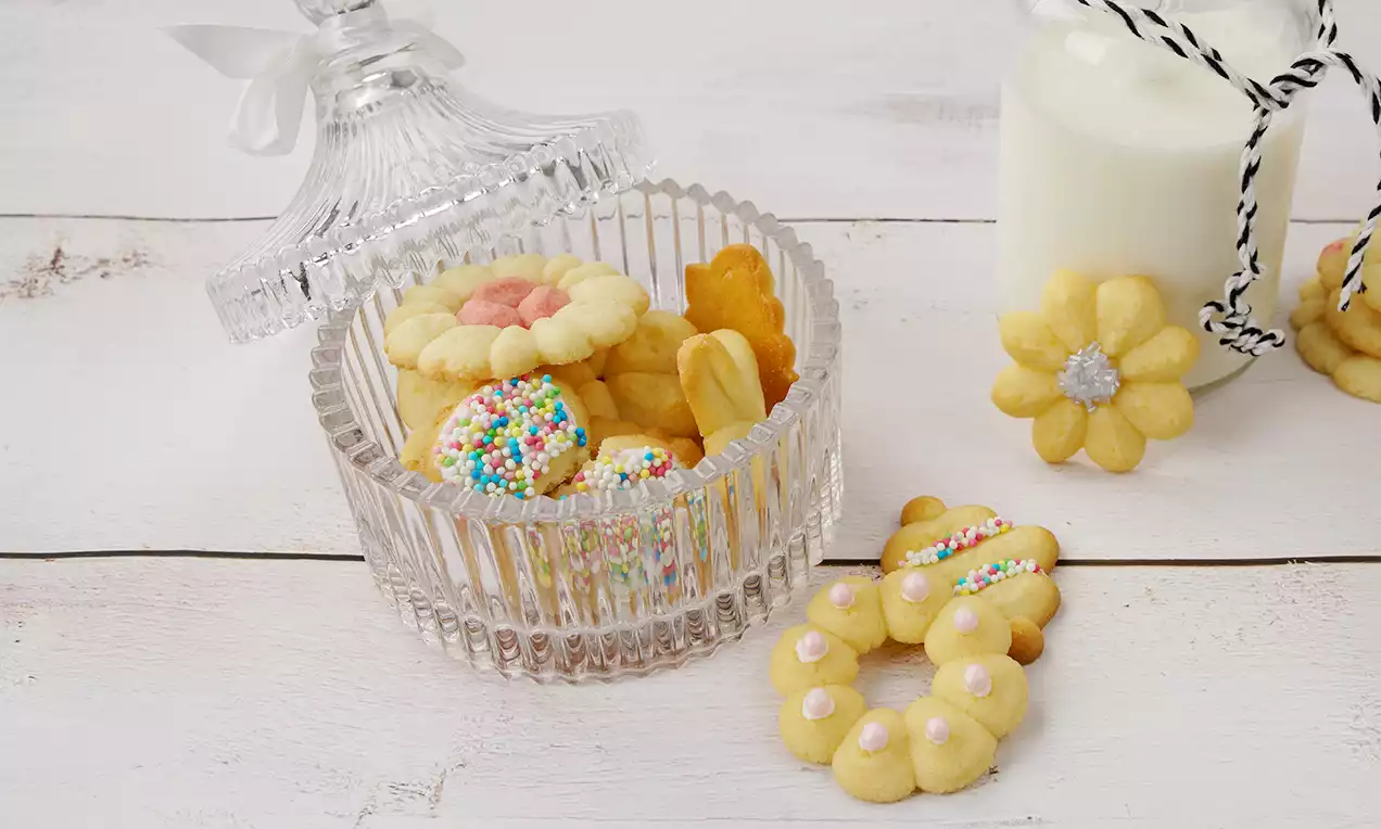 Buttergebäck in verschiedenen Formen, goldgelb und zart, teils mit bunten Streuseln auf einer facettierten Glasdose arrangiert