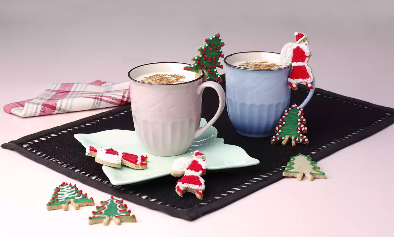 İki pastel renkli kupa, süslenmiş yılbaşı temalı kurabiyelerle birlikte siyah bir örtü üzerinde dekoratif şekilde sunulmuş