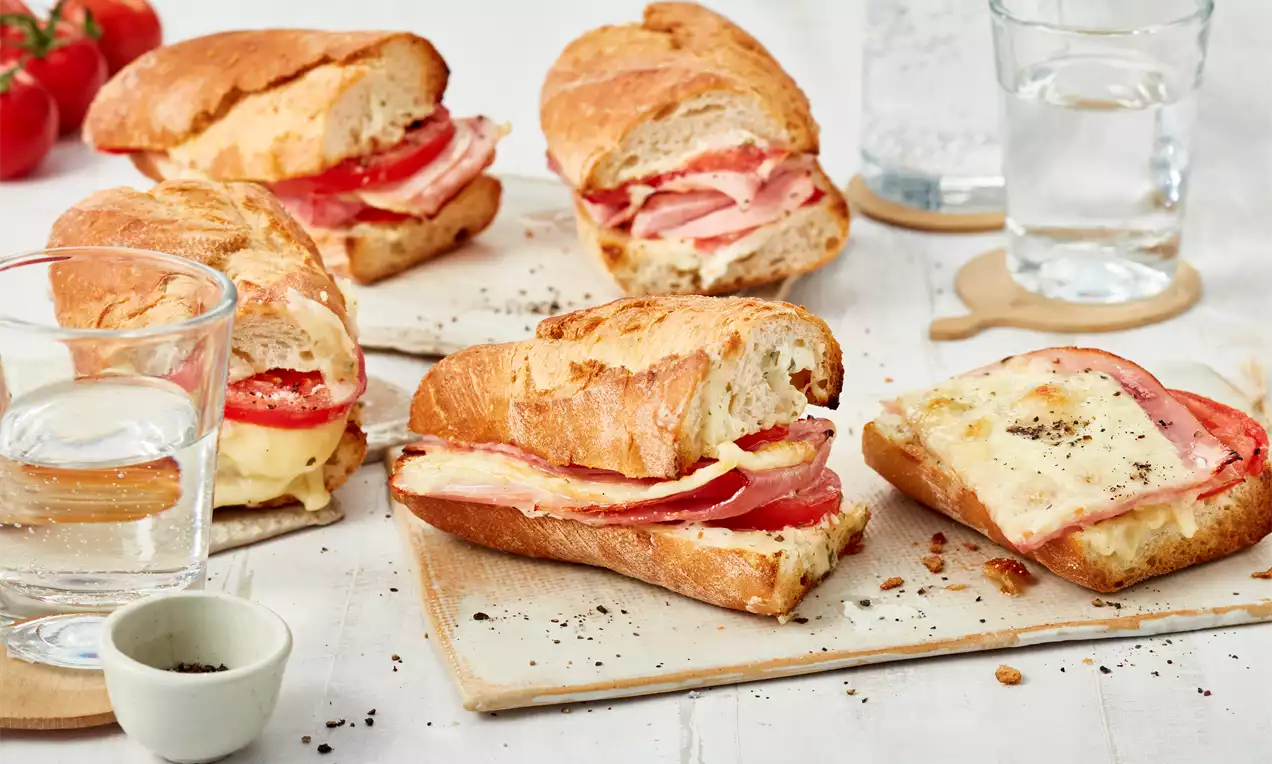 Knusprige Baguettehälften mit goldbraunem Käse, rosafarbenem Schinken und roten Tomatenscheiben auf hellem Holzbrett arrangiert