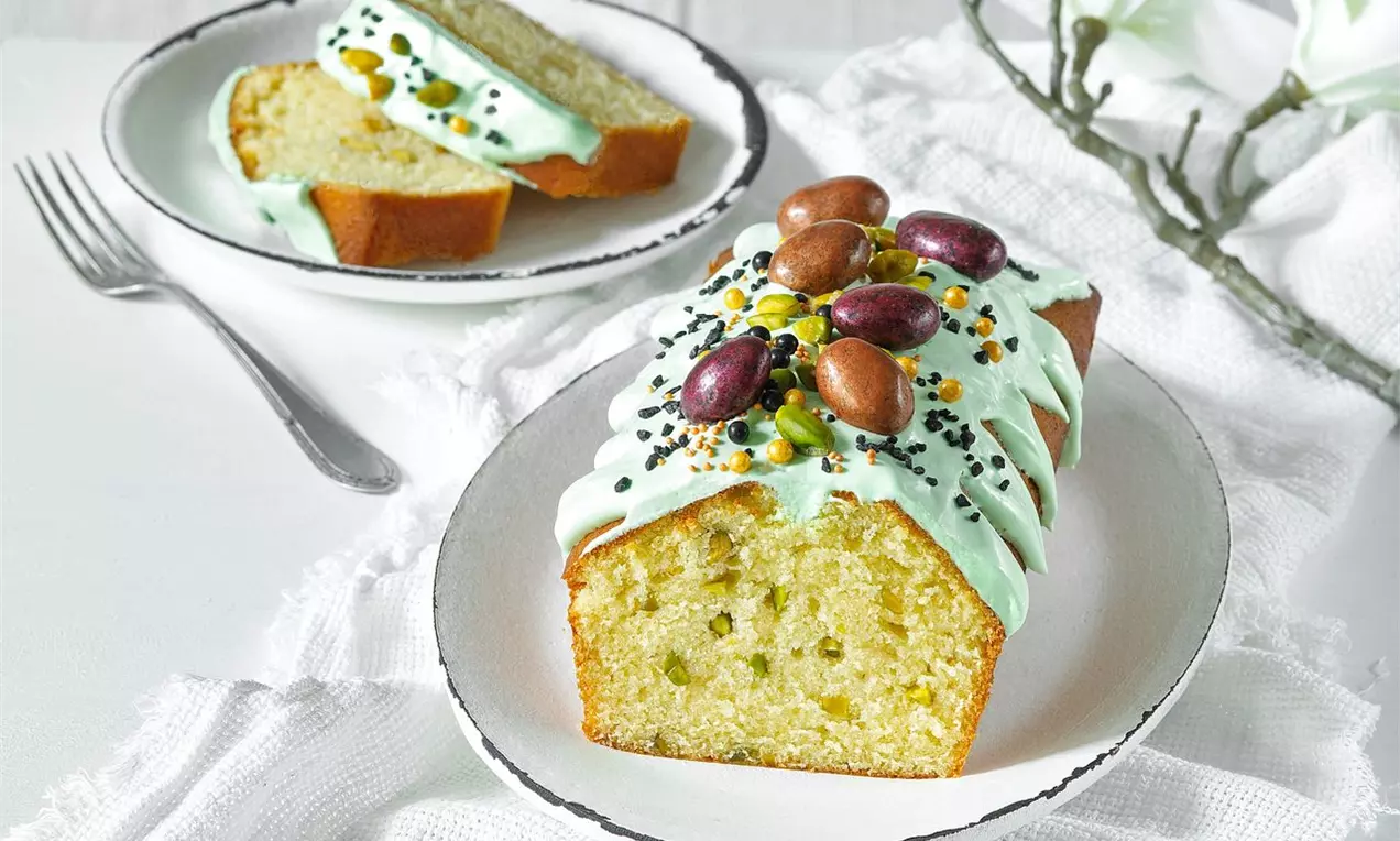 Veľkonočný chlebíček svetlozelený s pistáciami, poliaty krémovou polevou, zdobený farebnými vajíčkami a semienkami na bielom tanieri