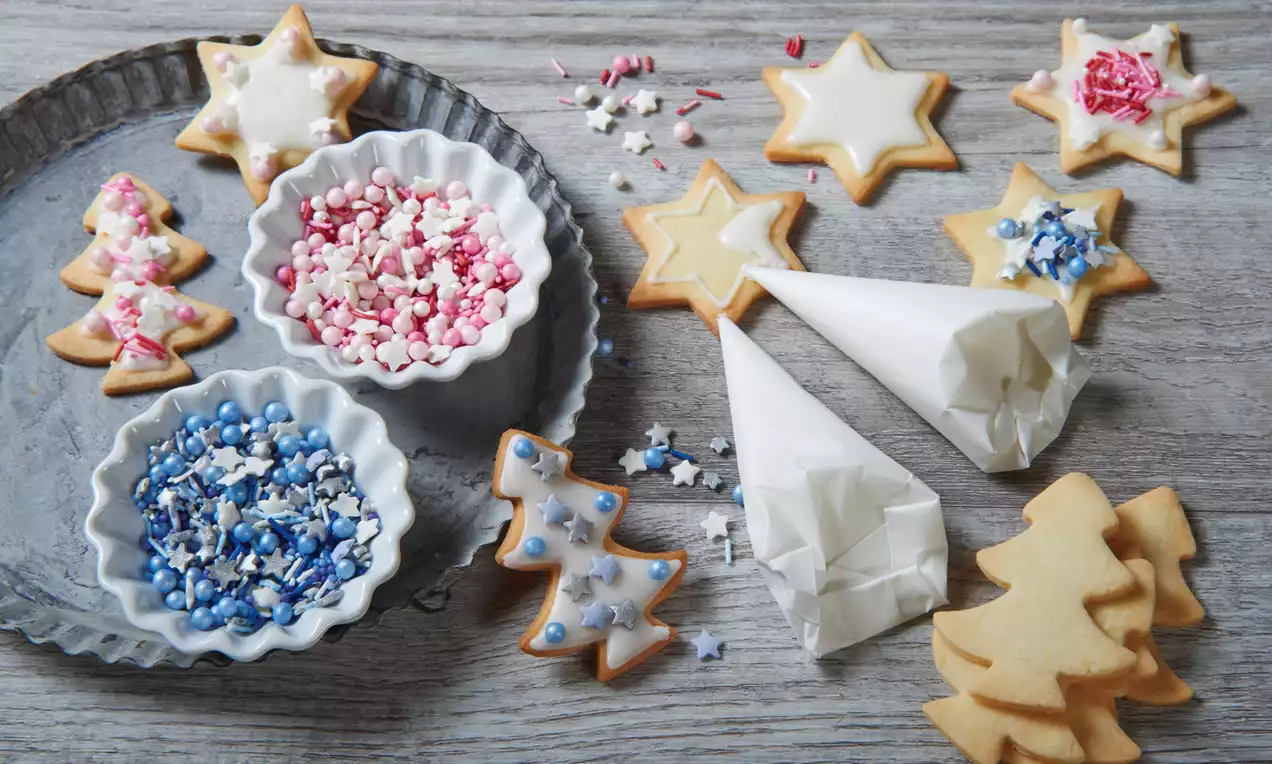 Weihnachtsplätzchen in Stern- und Tannenbaumform mit weißem Zuckerguss, farbigen Streuseln und Spritztüten auf grauem Holz und Metalltablett